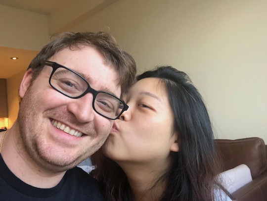 Yuankun kissing Ryan on the cheek while they smile on the couch.